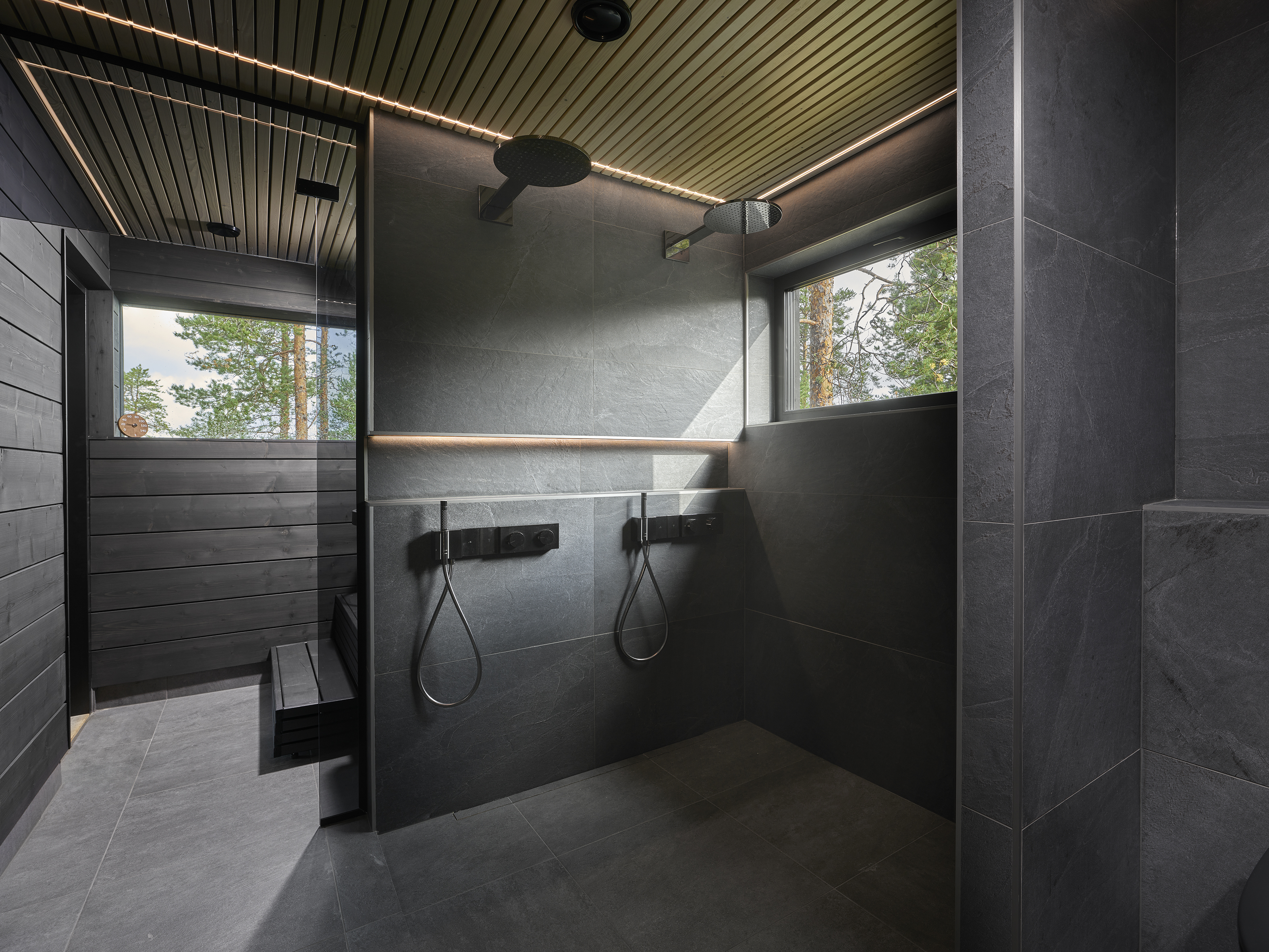 Dark-tiled bathroom with twin showers and a window onto the forest