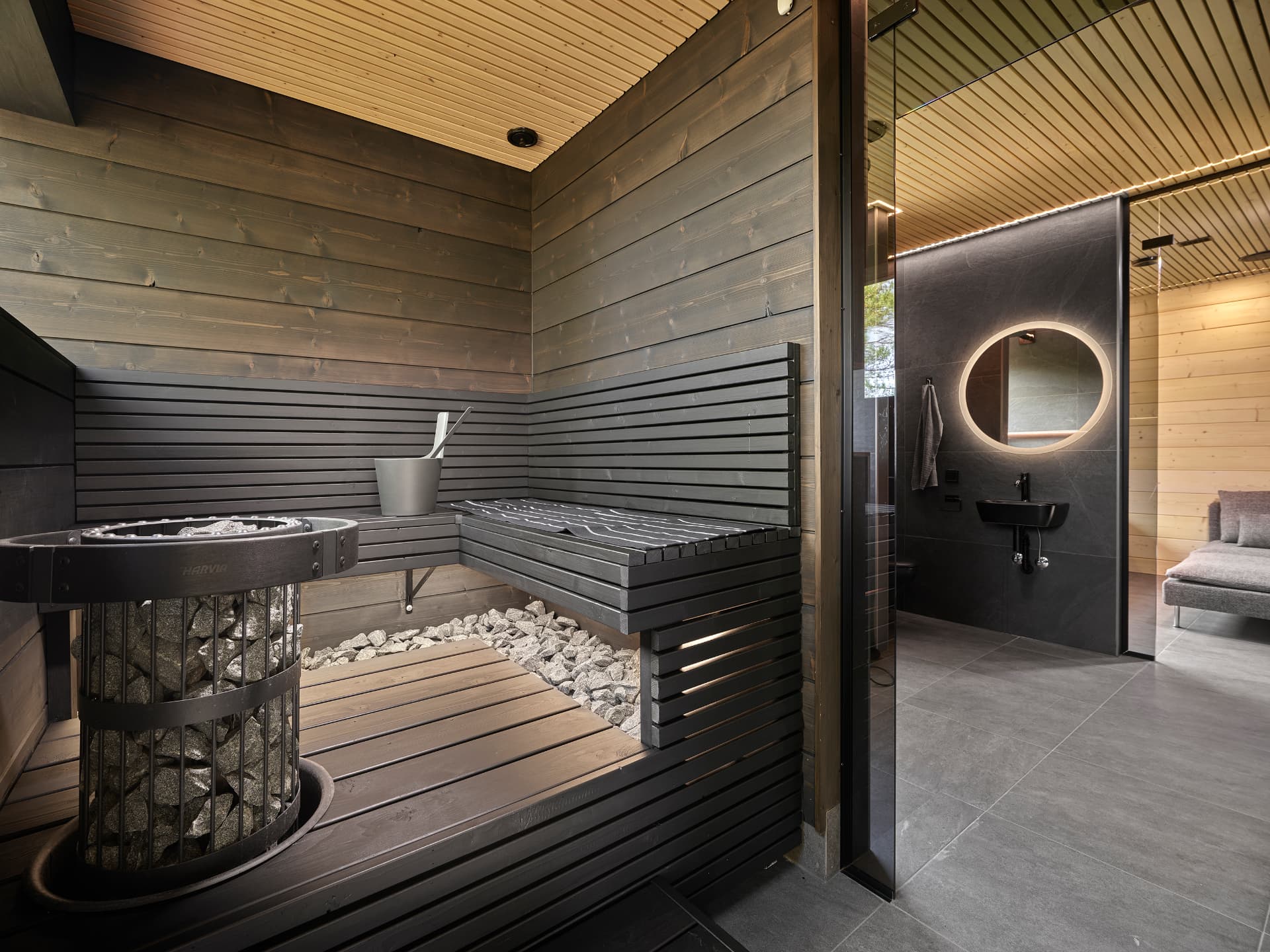 Sauna with dark timber benches, a stone-filled stove and warm bench lighting