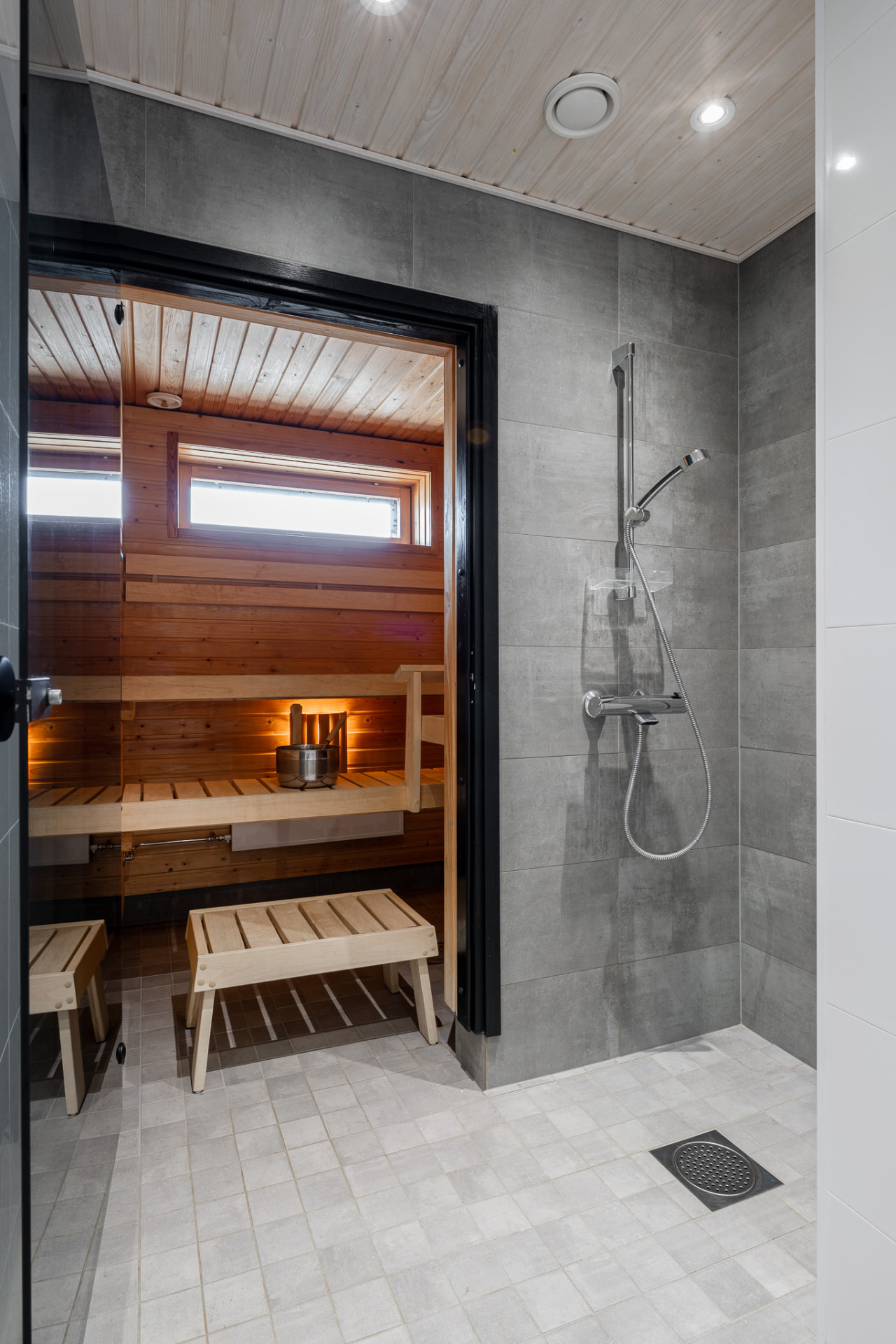 Walk-in shower with grey stone tiles beside the glass door to the warm wooden sauna