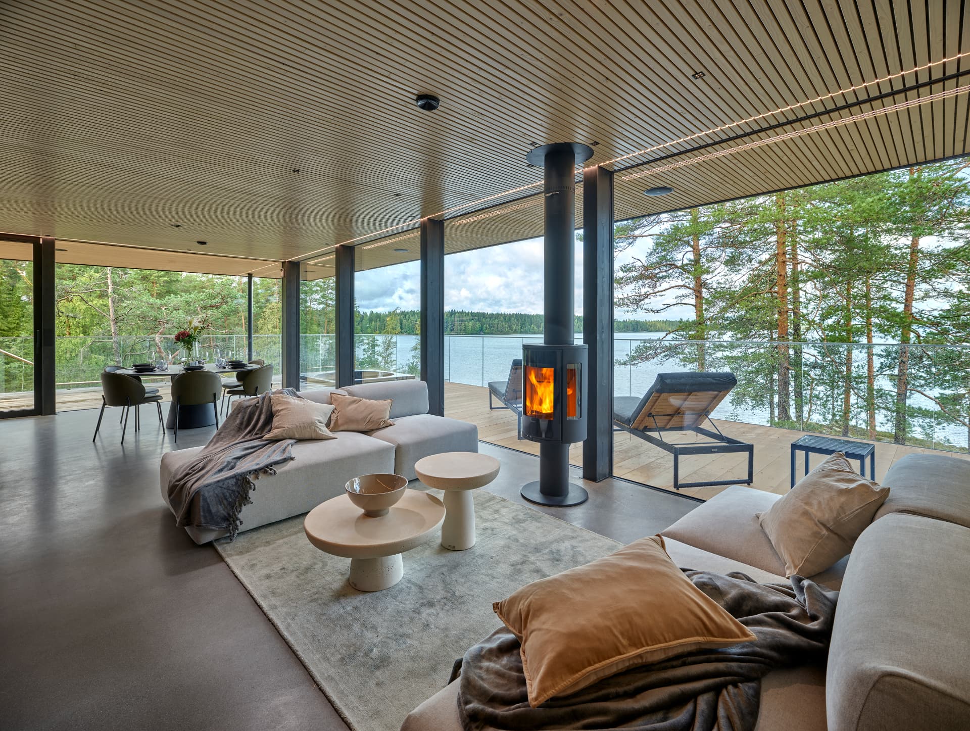 Living room with a wood-burning fireplace, soft neutral sofas and panoramic windows onto the lake and pines