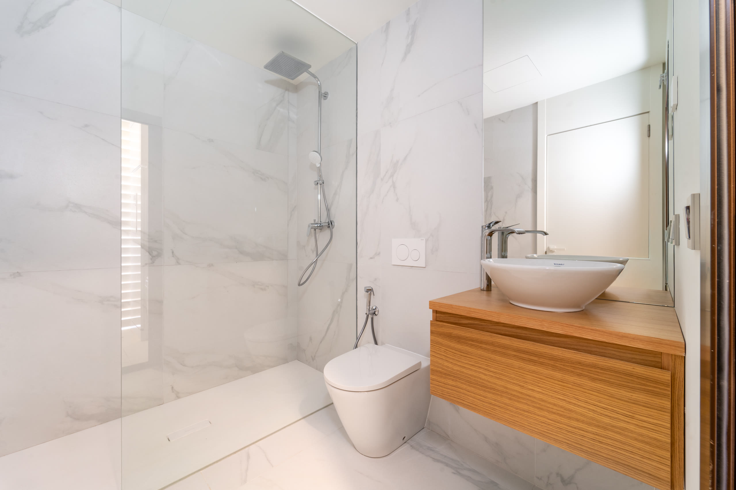 Walk-in shower in pale marble with warm wood vanity